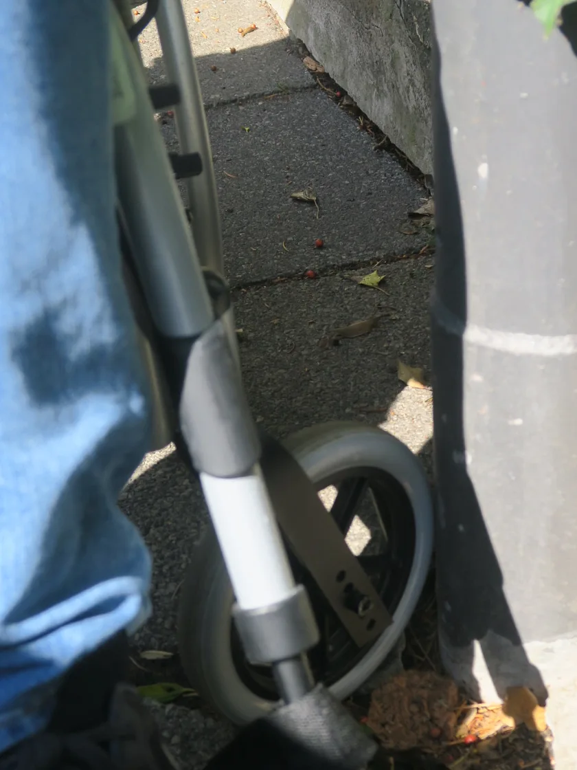 Das linke Vorderrad eines Rollstuhls hat sich in einem Loch einer Gehwegplatte an einem Laternenmast verkantet.