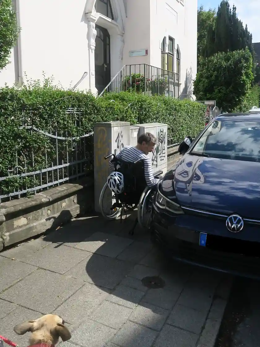Rollstuhlfaher beim passieren einer Engstelle, links vom Rollstuhlfahrer stehen Stromschaltk&auml;sten auf dem Gehweg, rechts parkt aufgesetzt ein VW